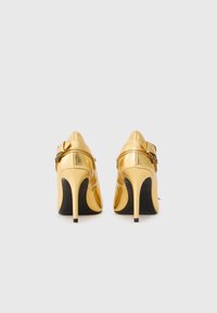 Gold high-heeled shoes with pointed toes, a smooth metallic finish, and a decorative strap with embellishments on the back.