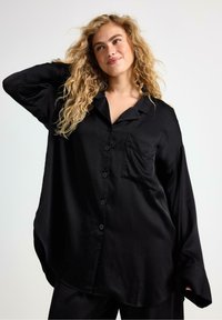 Blond kvinde med krøllet hår iført en oversized sort skjorte med knapper og lomme foran, som smiler og poserer med armen bag hovedet mod en hvid baggrund.