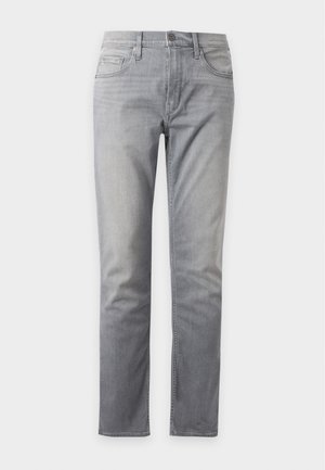 Jeune homme portant un polo manches longues vert olive, un jean gris clair et des mocassins marron foncé, debout avec une main dans la poche.