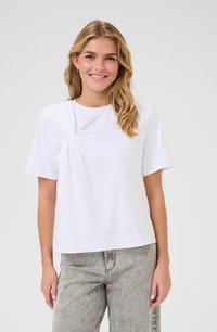 T-shirt blanc à manches courtes avec un détail torsadé sur le côté gauche, fabriqué en coton doux, associé à un jean taille haute gris.