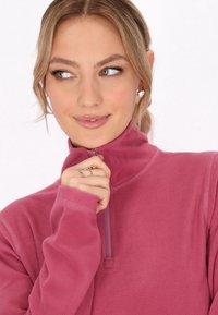 Fleece pullover in een levendige roze kleur, met een hoge kraag en een rits, lange mouwen en een zachte textuur. Hand rustend bij de kraag.