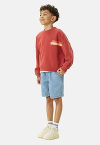 Sweat-shirt en coton rouge avec des graphismes blancs et orange, associé à un short en denim bleu avec un motif de sourire. Porté avec des baskets blanches.