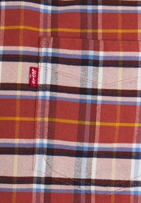 Camisa de cuadro rojo con acentos blancos, amarillos y azules. Fabricada en tejido de punto con un bolsillo en el pecho y una etiqueta de marca visible.