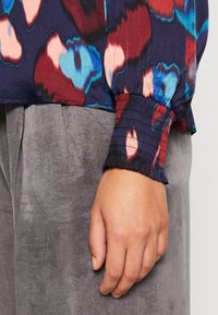 Main reposant sur le côté, portant un chemisier multicolore à manches longues avec poignet élastique et un pantalon gris texturé.