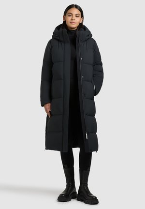 Lungo cappotto piumino nero con cappuccio, dotato di sezioni cucite, bottoni a pressione e zip frontale. Indossato sopra un completo nero e stivali.