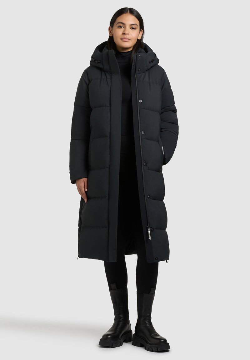 Langer schwarzer Daunenmantel mit Kapuze, der über genähte Abschnitte, Druckknöpfe und einen Frontreißverschluss verfügt. Getragen über einem schwarzen Outfit und Stiefeln.