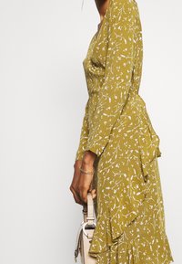 Sido vy av en person klädd i en olivgrön blommig klänning med volanger, som håller en beige texturerad handväska och bär ett guldarmbandskedja.