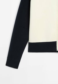 Manica lunga nera che si estende dal lato sinistro di un indumento color crema con un orlo nero nella parte inferiore.