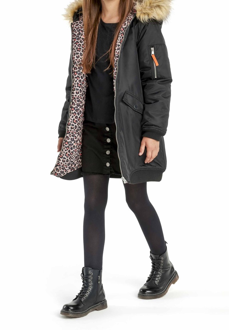 Enfant portant un parka noir avec capuche en fourrure et doublure imprimé léopard, jupe noire avec boutons, collants noirs et bottes noires à lacets en train de marcher.