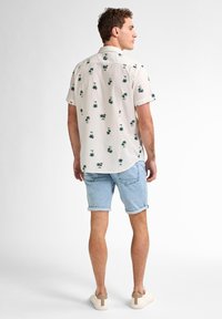 Camicia a maniche corte con bottoni, con sfondo bianco e motivi tropicali verdi. Abbinata a pantaloni corti in denim azzurro chiaro e scarpe da ginnastica beige.