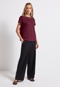 Bordeaux kortærmet bluse med en nøglehulldetalje, parret med løse sorte bukser og sorte mesh slip-on sko, stående imod en hvid baggrund.