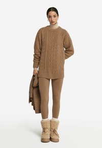Kabelstrickpullover in Hellbraun, kombiniert mit passenden Leggings und Schaffellstiefeln. Weiche Texturen und geschichtete Styling-Elemente sind besonders ausgeprägt.
