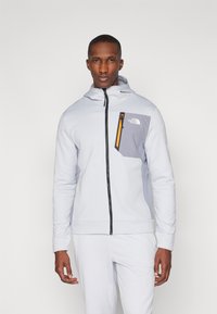 Lys grå zip-hoodie laget av myk stoff, med en kontrasterende grå lomme og en oransje glidelåsdetalj på brystet.
