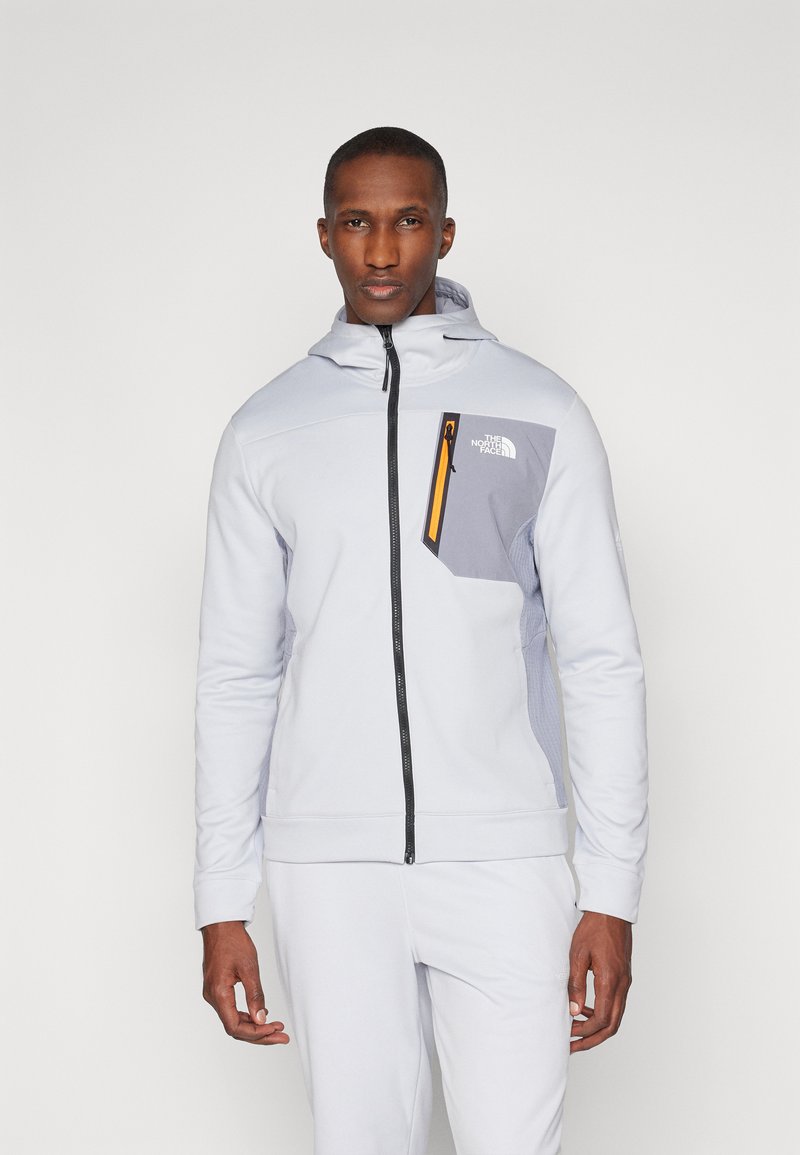 Lys grå zip-hoodie laget av myk stoff, med en kontrasterende grå lomme og en oransje glidelåsdetalj på brystet.