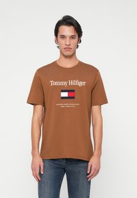 Rjava bombažna majica s krožnim ovratnikom, ki vsebuje logotip "Tommy Hilfiger", majhno grafiko zastave in besedilo "USTANOVLJENO MCMLXXXV NEW YORK CITY".