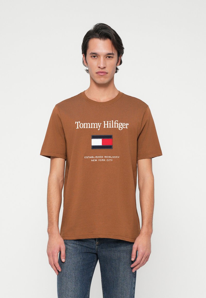 Rjava bombažna majica s krožnim ovratnikom, ki vsebuje logotip "Tommy Hilfiger", majhno grafiko zastave in besedilo "USTANOVLJENO MCMLXXXV NEW YORK CITY".