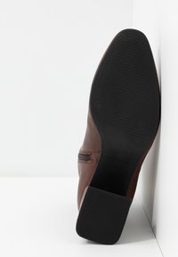 Botte en cuir marron avec semelle en caoutchouc noire et talon bloc, positionnée debout contre un mur d'angle blanc et un sol blanc.