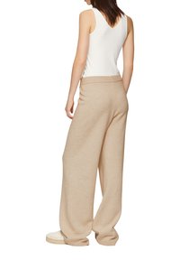 Beige wijde broek van een zachte, gestructureerde brei, met een ribbelband. Model draagt een witte tanktop en casual schoenen.