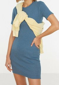 Kvinna som bär en figursydd kortärmad blå klänning med en blekgul tröja knuten runt axlarna och handen på höften.