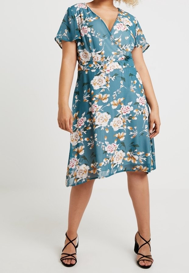 Femme portant une robe portefeuille turquoise longueur genou avec des imprimés floraux blancs et roses, et des sandales à talons noires à brides.