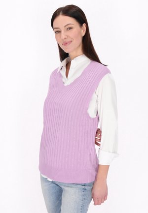 Kvinde med langt mørkt hår iført en hvid skjorte og en ærmeløs lavendel kabelstrikket sweater, stående mod en ensfarvet hvid baggrund.