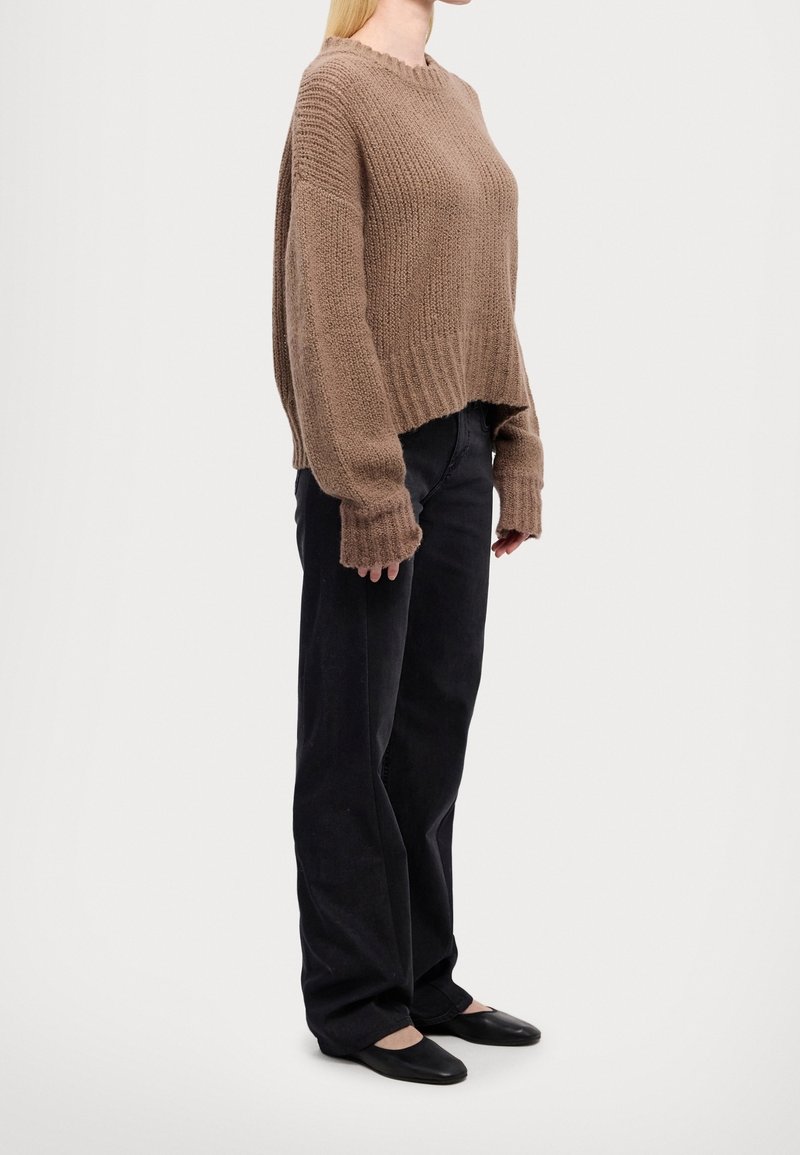 Personne portant un pull en tricot marron ample, un pantalon noir à jambes larges et des ballerines noires, debout devant un fond blanc uni.