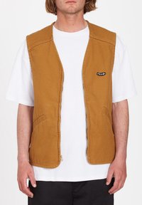 Homme portant une veste sans manches marron zippée sur un t-shirt blanc, debout devant un fond blanc uni.