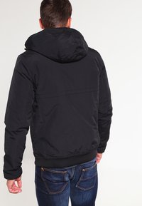 Giacca nera con cappuccio, realizzata in un tessuto morbido, con polsini elastici e vita a coste, indossata sopra un paio di jeans blu. Presenta un design semplice e lineare.