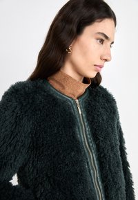 Groene nepbontjas met een ritssluiting aan de voorkant, lange mouwen en een gladde leren afwerking op de kraag. Gedragen over een bruine glinsterende coltrui.