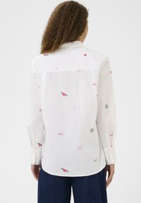 Göndör hajú nő fehér, hosszú ujjú inget visel, amelyen kis piros és rózsaszín szív, virág és "oui" feliratminták találhatók, sötétkék nadrággal kombinálva.