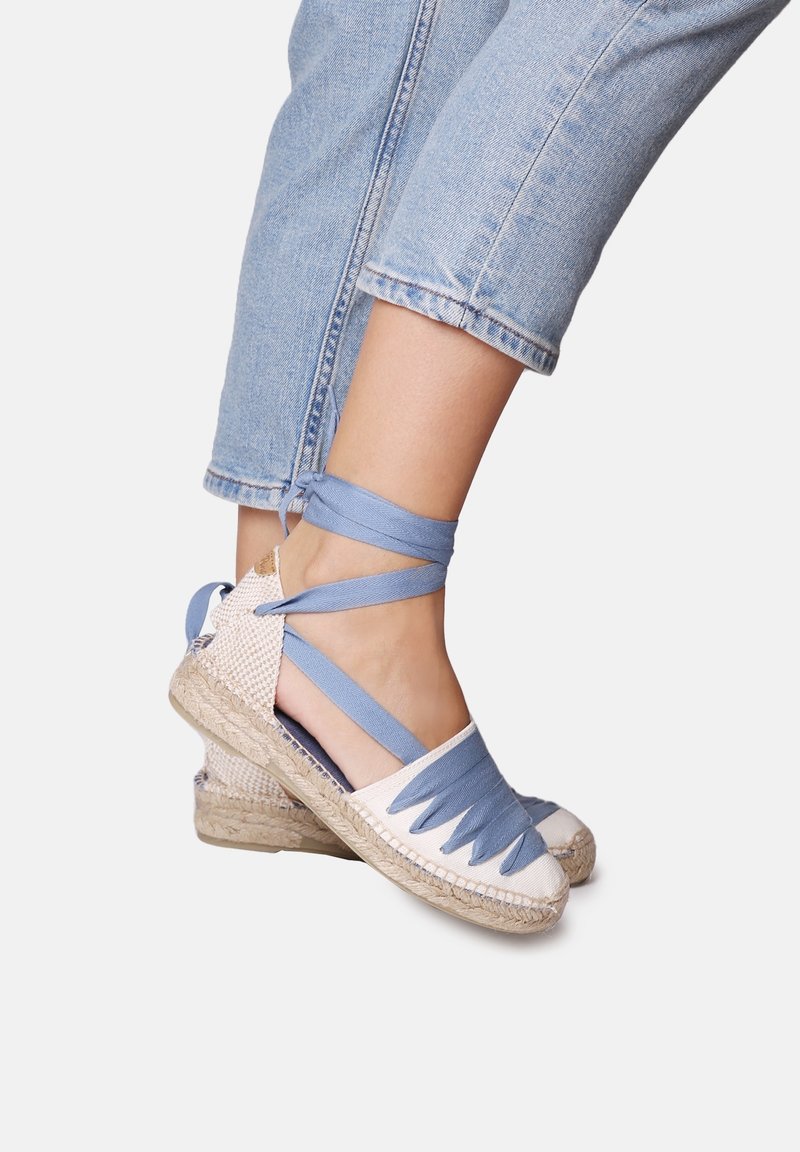 Porte des compensées espadrille bleu clair avec semelles en jute tissée et jean en denim bleu clair retroussé, sur un fond uni.
