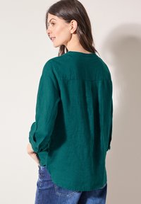 Türkisfarbenes Leinenhemd mit hochgekrempelten Ärmeln, einer Rückenpasse und einem abgerundeten Saum. Kombiniert mit blauen Jeans.
