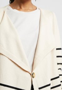 Beżowy kardigan z szerokim kołnierzem, z czarnymi paskami na rękawach i zapięciem na guziki z przodu. Miękka tekstura, casualowy design.