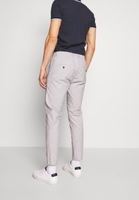 Muž v světle šedých slim kalhotách, bílých teniskách, bílých ponožkách a námořnickém tričku s krátkým rukávem stojící na jednobarevném pozadí.