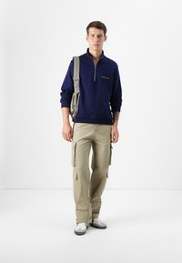 Felpa con zip blu marina con logo, abbinata a pantaloni cargo beige chiaro con diverse tasche. Borsa a spalla grigia e sneakers bianche.