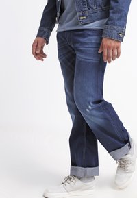 Jeans in denim blu scuro con effetto sbiadito sulle cosce, con risvolti arrotolati e tasche laterali, abbinati a sneakers bianche.
