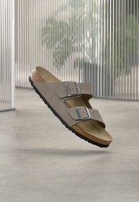 Μπεζ σανδάλι Birkenstock με δύο ρυθμιζόμενες ασημένιες αγκράφες και πάτο από φελλό, αιωρούμενο πάνω από τσιμεντένιο πάτωμα με θολό φόντο φυτού.