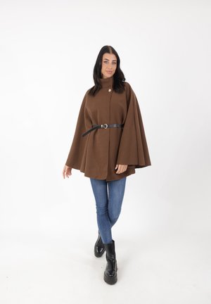 Cappotto marrone in stile mantella con colletto alto, chiusura frontale a bottone e un dettaglio in cintura nera. Abbinato a jeans blu e stivaletti neri.
