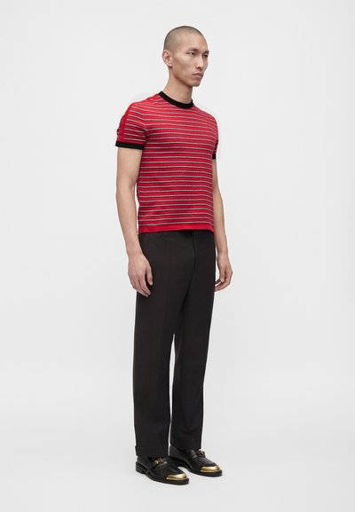 Homme debout portant une chemise à manches courtes rayée rouge et noire, un pantalon noir et des chaussures noires avec des embouts dorés sur fond blanc.