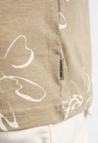 Short-sleeve beige T-shirt with white abstract print, featuring a stitched hem and a small branded label on the side.