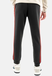 Pantaloni da jogger neri con strisce laterali rosse, vestibilità a sigaretta, tessuto morbido e polsini a costine. Indossati con scarpe bianche arricchite da accenti grigi e blu navy.