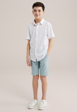 Vit kortärmad skjorta med knappar, tillsammans med ljusblå shorts. Outfiten fulländas med vita sneakers med ljusblå detaljer.