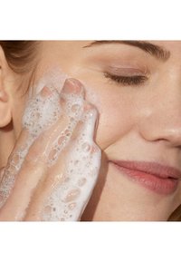 Ansiktsrens med rik, hvit skum påført huden. Nærbilde av en hånd som forsiktig masserer skummet på en glatt, lys hudtone.