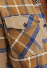 Bruine geruite shirt met een patchetas, blauwe en witte lijnen, en zwarte knopen. Het materiaal lijkt zacht en textuur te hebben met een flanellen uitstraling.