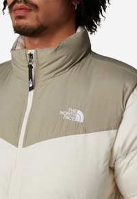 Puffjacka i ljusbeige och cream, med hög krage, dragkedjestängning och broderad logotyp på bröstet. Slät textur och stilren design.