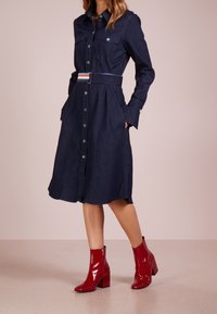 Tummansininen farkkupaitamekko, jossa napit, kaksi rintataskua ja vyötäröllä vyö. Yhdistetty kiiltäviin punaisiin nilkkureihin. Sileä pinta.