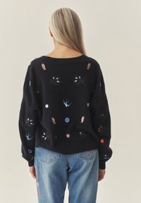 Czarny sweter wykonany z miękkiego materiału, zdobiony kolorową haftowaną grafiką postaci, kwiatów i kółek, o luźnym kroju i szerokich rękawach.