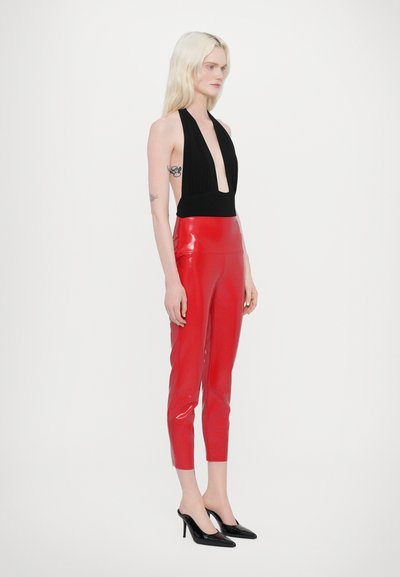 Røde, skinnende latexbukser med høj talje og slim fit, parret med en sort top med dyb V-udskæring. Modellen bærer sorte spidse hæle.