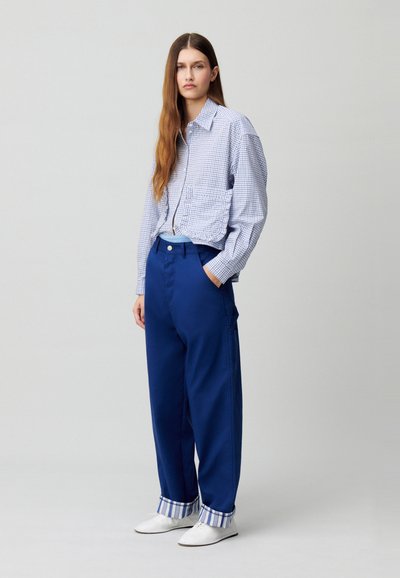 Camicia a quadri blu con vestibilità ampia e due tasche sul petto, abbinata a pantaloni blu larghi con polsini a righe e scarpe bianche.