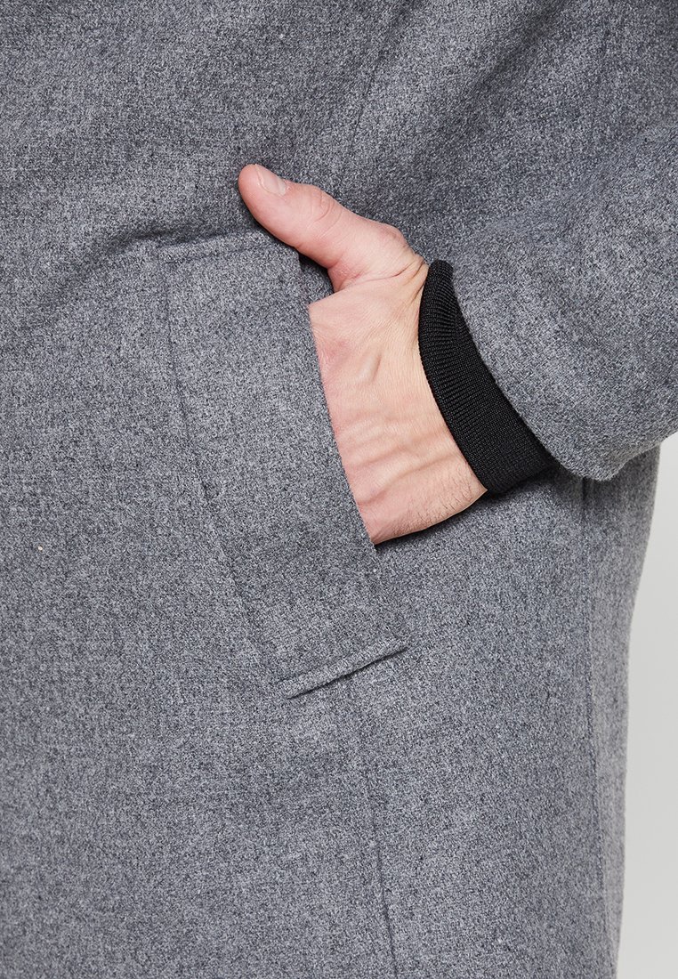 Szara polarowa kurtka z ręką częściowo włożoną w boczną kieszeń. Kieszeń charakteryzuje się schludnym, zorganizowanym wzornictwem oraz gładką teksturą.